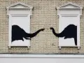 Œuvre Banksy : deux éléphants noirs se touchent la trompe à travers deux fenêtres sur un mur de briques.