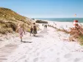 Familj promenerar på en strand med vit sand i Danmark. En livräddartorn och bunkrar syns i bakgrunden.