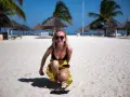 Uśmiechnięta blondynka w bikini i żółtej spódniczce kuca na piaszczystej plaży na Zanzibarze. W tle palmy i słomiane parasole.