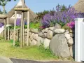 Natürliche Steingarten Ideen: Eine Steinmauer mit Lavendel und rosa Blüten, flankiert von jungen Bäumen und einem Reetdachhaus.
