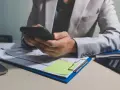 Osoba w jasnej marynarce przegląda telefon, na biurku dokumenty z wykresem i notatkę z informacją o uchyleniu zajęcia.