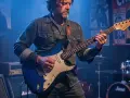 Un guitariste joue sur scène, évoquant l'esprit de qui a inventé le blues. Des affiches de concerts et des amplis Marshall créent une ambiance rock.
