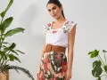 Mujer con top blanco bordado y falda plisada de estampado abstracto. Un cinturón marrón ancho realza la cintura.