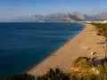 Słoneczna plaża na riwierze tureckiej z turkusowym morzem i górami w tle. Kilka osób pływa, inni odpoczywają na piasku.