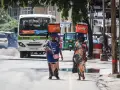 Dwie kobiety niosą na głowach pomarańczowe lodówki w upalny dzień w Dar es Salaam. W tle autobus i ruchliwa ulica.