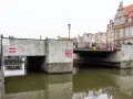 Nad kanałem w Gdańsku wznosi się betonowy zielony most, ozdobiony czerwono-białymi znakami. Ludzie spacerują po jego balustradzie.