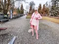 Kobieta w pudrowym różu, w płaszczu, sukience i kozakach, spaceruje po parku.