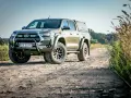 Złota toyota terenowa z wyciągarką i zabudową pędzi po polnej drodze, otoczona dziką roślinnością.