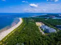 Widok z lotu ptaka na najszerszą plażę w Polsce, pełną wypoczywających ludzi, otoczoną lasem i nowoczesnymi budynkami.