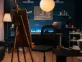 Un estudio de pintura en casa con caballete, silla de oficina, lámpara de pie y un escritorio con ordenador.