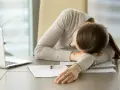 Kobieta z opuszczoną głową na rękach przy biurku, obok laptopa i dokumentów. Widać wyraźny brak motywacji do czegokolwiek.