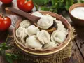 Pielmieni co to? Pyszne, gotowane pierożki w glinianej misce, z drewnianą łyżką, pomidorami i śmietaną w tle.
