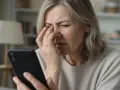 Donna anziana con disturbi visivi, forse cerchi luminosi, si tocca gli occhi mentre guarda uno smartphone.