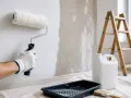 Mano guantata stuccare una parete prima di pitturare con rullo. Attrezzi da lavoro e scala sullo sfondo.