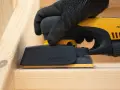 En hand med handske slipar en träyta med en Mirka DEOS-slipmaskin. Perfekt för att slipa borr och andra trädetaljer.