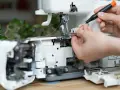 Dłonie regulują maszynę do szycia, używając śrubokręta. Widać fragmenty mechanizmu i nitki, a także pasek napędowy.