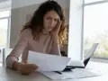 Kobieta zmartwiona przegląda dokumenty przy biurku z laptopem.