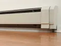 Un zócalo radiante blanco instalado en una pared, con el suelo de madera. Sus desventajas pueden incluir la estética o la distribución del calor.