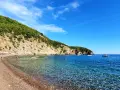 Słoneczne plaże w Budvie, z turkusową wodą i zielonymi wzgórzami w tle. Ludzie relaksują się na kamienistym brzegu i pływają w morzu.