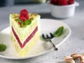 Pyszny tort pistacjowy z malinowym nadzieniem, udekorowany malinami i miętą. Obok leżą łuskane pistacje.
