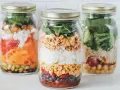 Drei bunte Schichtsalate im Glas, bereit für unterwegs. Jedes Glas ist ein kleines Kunstwerk mit Reis, Gemüse und Proteinen.