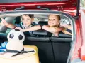 Två glada barn i bilens bagageutrymme, redo för äventyr. En fotboll och en hatt ligger redo, en perfekt resa med barn packlista.