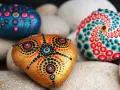 Drei bunte, bemalte Steine liegen auf hellen Kieselsteinen. Mandala-Muster in Rot, Blau und Pink zieren die Steine.