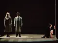 Magdalena Twardowska w scenie teatralnej. Dwie postacie w płaszczach stoją w świetle, obok nich kobieta w czarnej sukni klęczy przy pionowym świetle.