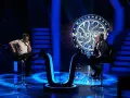 Dwóch mężczyzn siedzi naprzeciwko siebie na scenie "Milionerzy". Jeden w białej koszulce, drugi w garniturze.