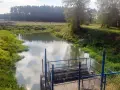 Spokojna rzeka otoczona zielenią, z widocznym pomostem i ogrodzeniem. Piękne zdjecia w wodzie odbijające niebo.