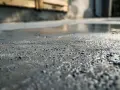 Zbliżenie na mokrą powierzchnię, gdzie trwa ręczne zacieranie betonu cementem. Widać drobne kamyczki i nierówności.