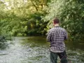 Mężczyzna w kratę łowi ryby w rzece. Woda sięga mu do pasa, a w ręku trzyma wędkę. W tle bujna zieleń drzew. To idealna przykosa na rzece.
