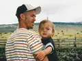 Ein Mann mit Bart und Kappe trägt ein Kind. Die Eigenschaften eines Papas zeigen sich in seiner liebevollen Umarmung.