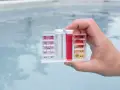 Hand holds pool water test kit. Red liquid in the pH tube indicates a high reading, suggesting acid is needed to lower pH.