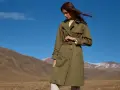 Mujer con un trench verde oliva, cinturón anudado, en un paisaje montañoso con cielo azul.