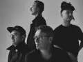 Czterech mężczyzn w czarnych ubraniach, spięty black metalowy zespół, z różnymi fryzurami i okularami, pozuje do zdjęcia.