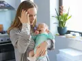 Müde Mutter hält weinendes Baby. Der **12 wochen schub** macht ihr zu schaffen. Sie hält sich die Stirn.