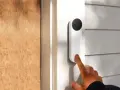 Un dedo presiona qué es el timbre inteligente, un dispositivo blanco con cámara negra, junto a una puerta de madera.