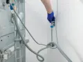 Blaue Handschuhe schließen den Wasserhahn für den Waschmaschinen Anschluss. Ein Schlauch führt zur Rückseite der Maschine.