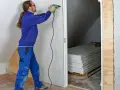 Man med skruvdragare monterar gipsskivor, kanske för att fixa en spackelremsa jula. Staplade skivor syns i bakgrunden.