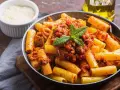 Un plato de macarrones con carne picada, cubiertos con salsa de tomate y decorados con menta. Al lado, un cuenco de queso rallado y una botella de aceite de oliva.