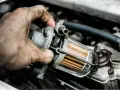 Brudna ręka mechanika wymienia filtr LPG w samochodzie. Widać przezroczysty element z pomarańczowymi wkładami.