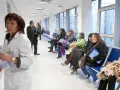 Doctora con bata blanca revisa papeles, mientras pacientes esperan en pasillo. Quizás pensando en cómo pedir excedencia estando de baja.