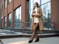 Elegancka kobieta w płaszczu w camelowy kolorze i brązowych spodniach ze skóry ekologicznej pozuje na tle nowoczesnego budynku.
