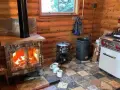 Poêle à bois allumé dans un chalet en bois rond, respectant la norme d'installation. Cuisine fonctionnelle avec cuisinière et ustensiles.