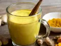 Taza de leche dorada con canela, rodeada de raíz de cúrcuma fresca y en polvo.