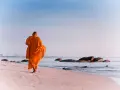 A modern day monk in an orange robe walks along a serene beach at sunrise, with calm ocean waves and distant city skyline.