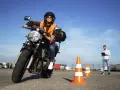 Joven sonriente en moto, lista para obtener el carnet de moto 49cc. Instructor observa en el fondo.