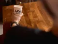 Dłoń trzyma kubek z logo "JAVA COFFEE ROASTERS", gotowa do degustacji świeżego batch brew na drewnianym stole.