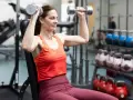 Kvinna utför axelpress med hantlar i gym. Hon sitter på en bänk och lyfter vikterna över huvudet.